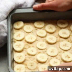Peanut Butter Banana Oatmeal Bake 4 Person placing sliced bananas onto banana baked oatmeal batter in a baking tray, with whole bananas and a cloth in the background.