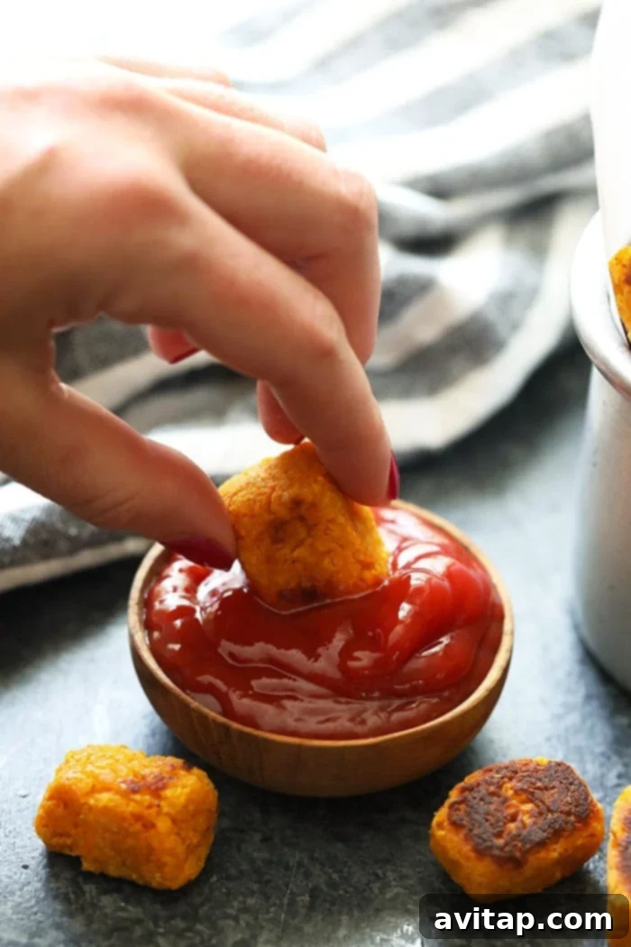 A hand dipping a crispy baked sweet potato tater tot into a small bowl of ketchup.
