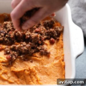 A person preparing a healthy sweet potato casserole by spreading mashed sweet potatoes into a dish and adding the streusel topping.