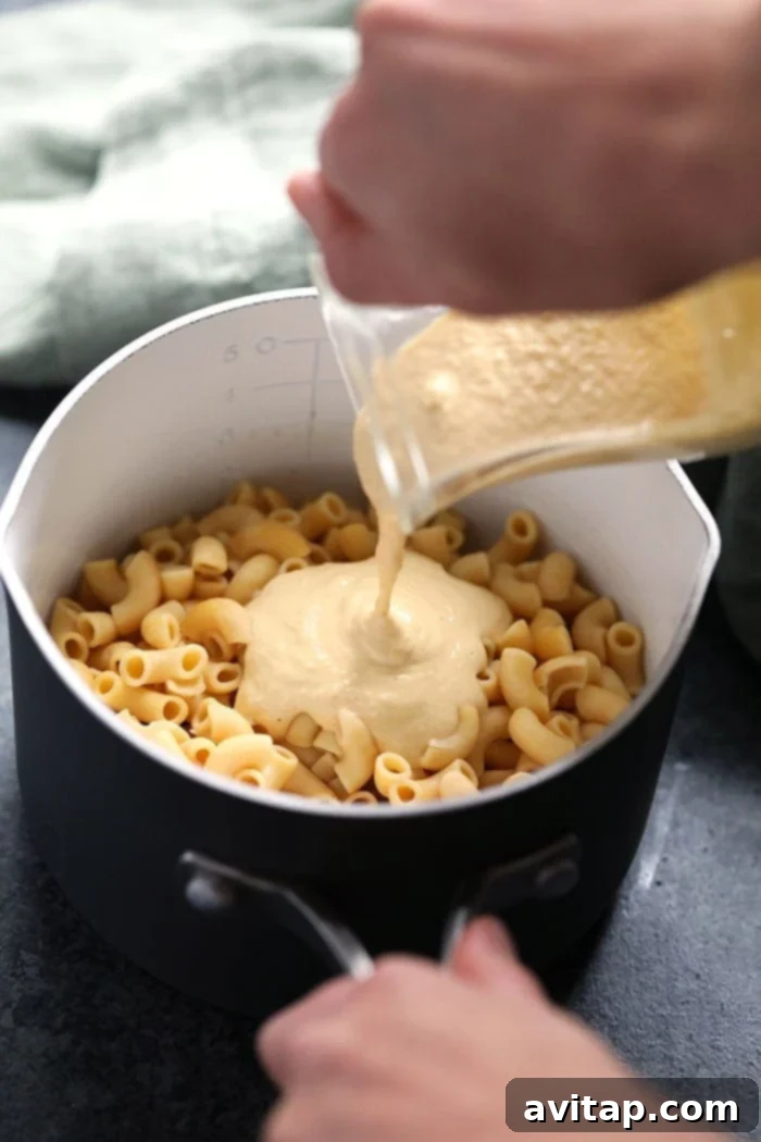 Rich vegan mac and cheese sauce being generously poured over a bed of perfectly cooked noodles in a large pan.