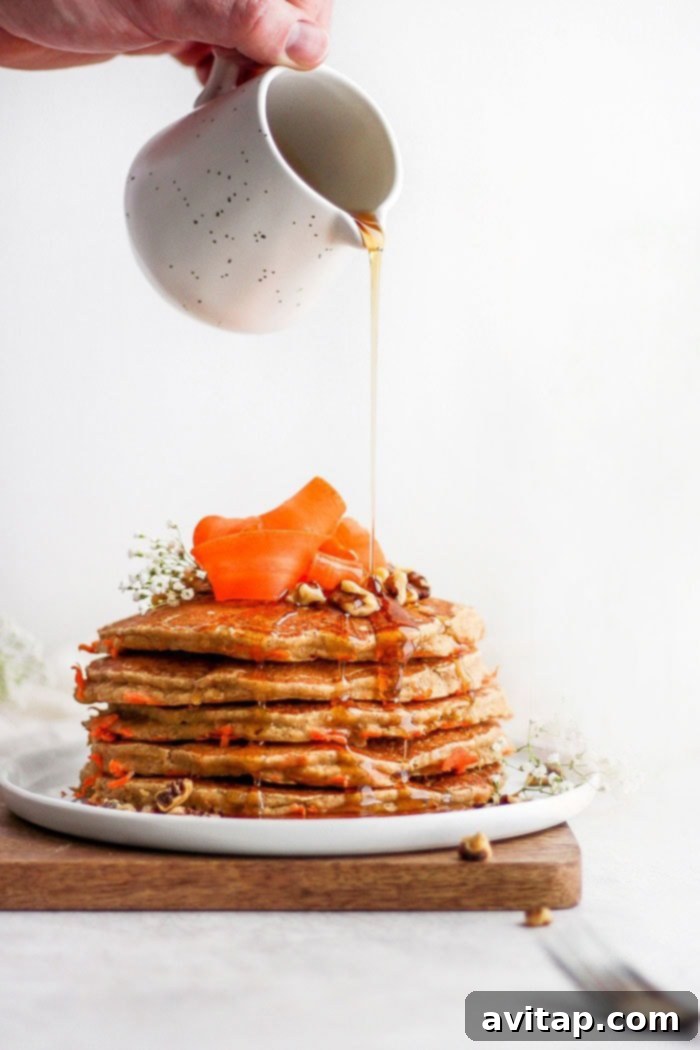 Stack of healthy carrot cake pancakes on a plate, garnished with maple syrup and nuts.