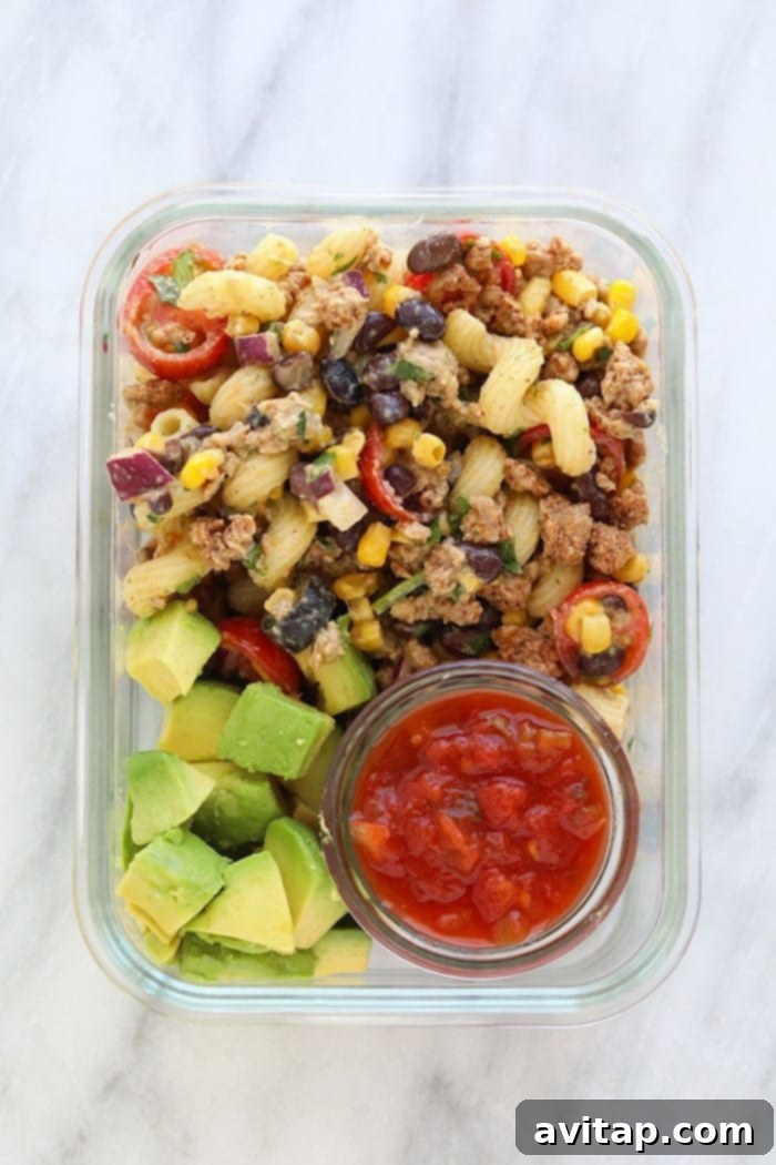 A perfectly portioned meal prep container featuring healthy taco pasta salad, accompanied by fresh sliced avocado and a separate side of salsa, ready for a delicious and convenient meal.