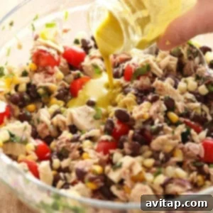 A hand pouring dressing into a glass bowl filled with a vibrant fiesta tuna salad, brimming with beans, corn, tomatoes, herbs, and chunks of tuna.