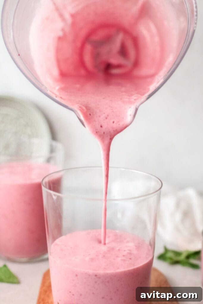 A creamy strawberry banana smoothie being poured from a blender into a tall glass, highlighting its rich, smooth texture and vibrant pink color.