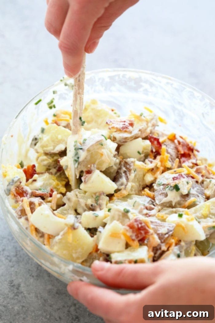 A hand gently mixing a large bowl of loaded baked potato salad, showcasing the creamy texture and generous add-ins.