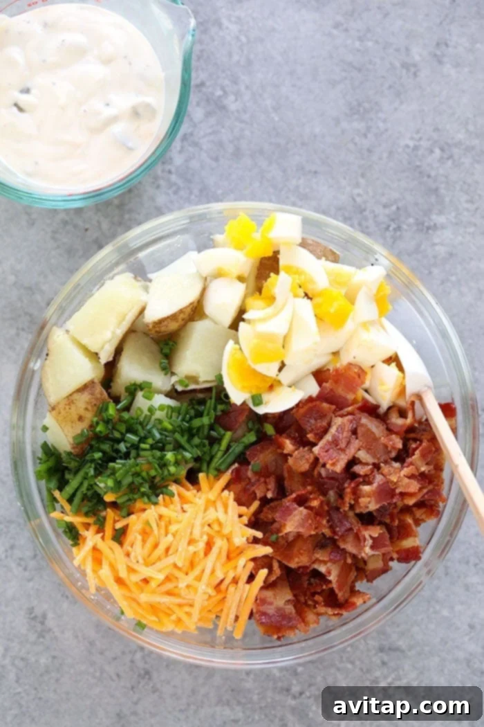 All the fresh ingredients laid out for making loaded potato salad: potatoes, bacon, cheese, chives, eggs, and sauce components.