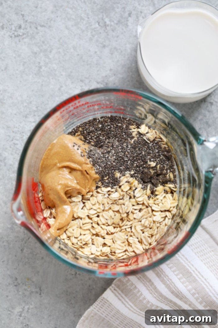 A close-up shot of the core ingredients for Peanut Butter Overnight Oats: oats, a spoon of peanut butter, a small bowl of chia seeds, and a glass of almond milk.