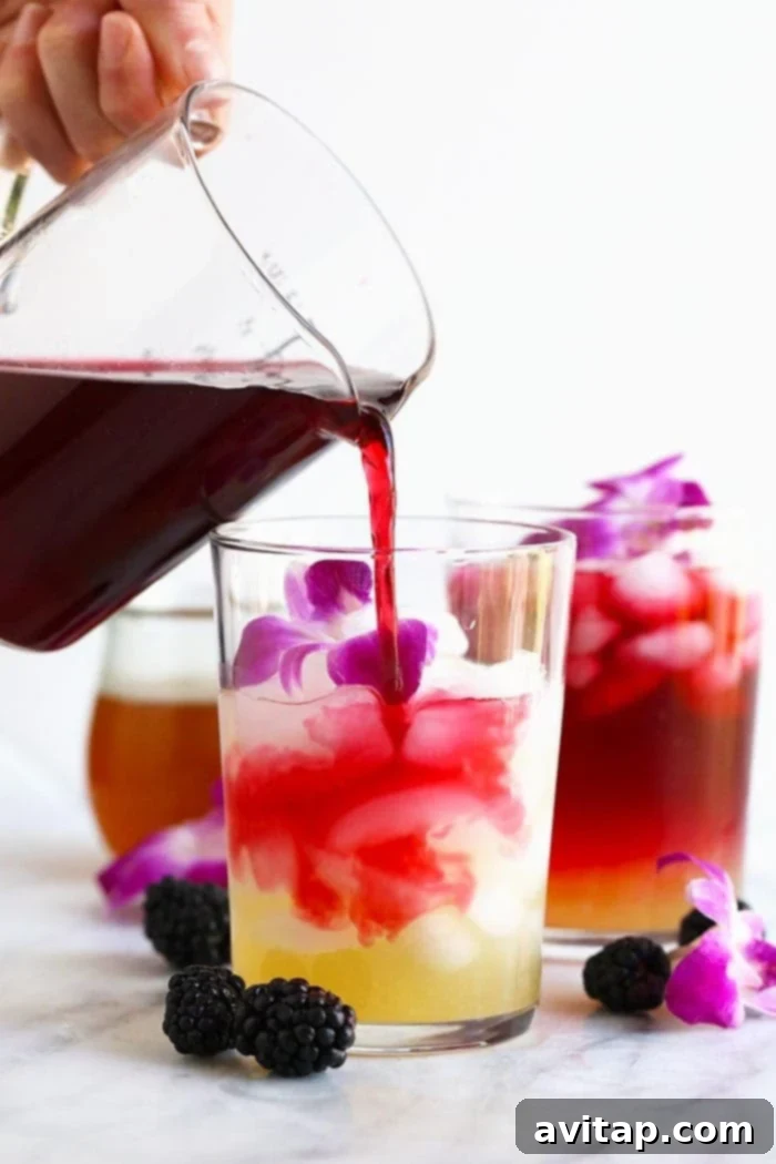 A vibrant Hibiscus Arnold Palmer drink in a clear glass, beautifully garnished with delicate edible flowers and a wedge of fresh lemon. The drink boasts a striking deep pink hue.