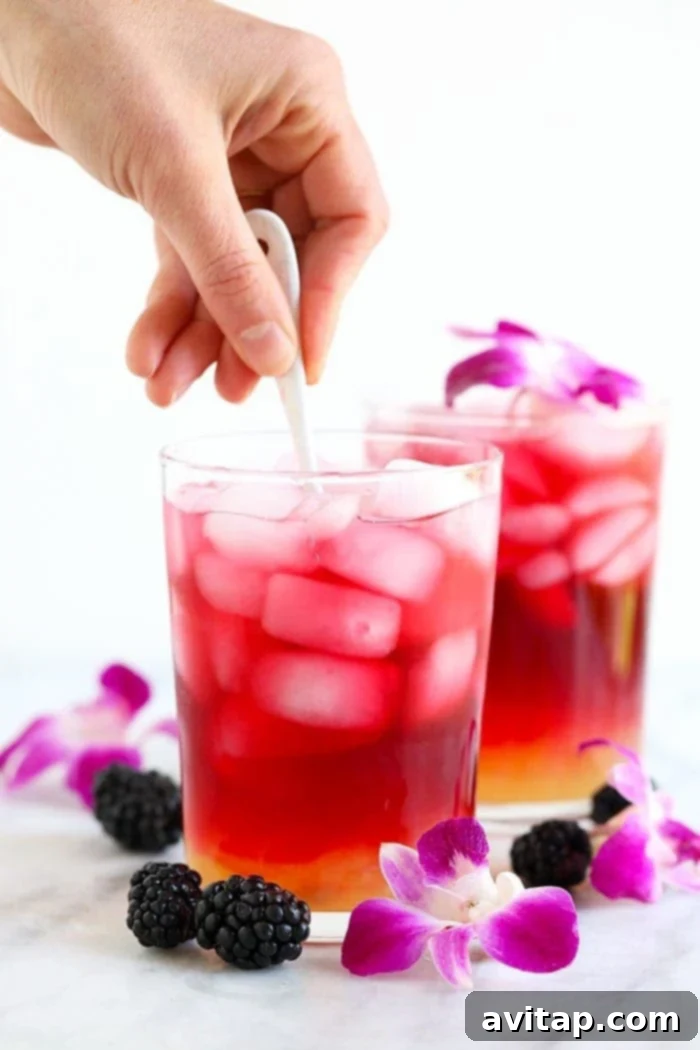 A clear glass showcasing a beautiful, swirling Hibiscus Arnold Palmer drink, being gently stirred with a long spoon. The distinct red-pink hue of the hibiscus tea merges gracefully with the lighter yellow of the lemonade, indicating a perfect blend.