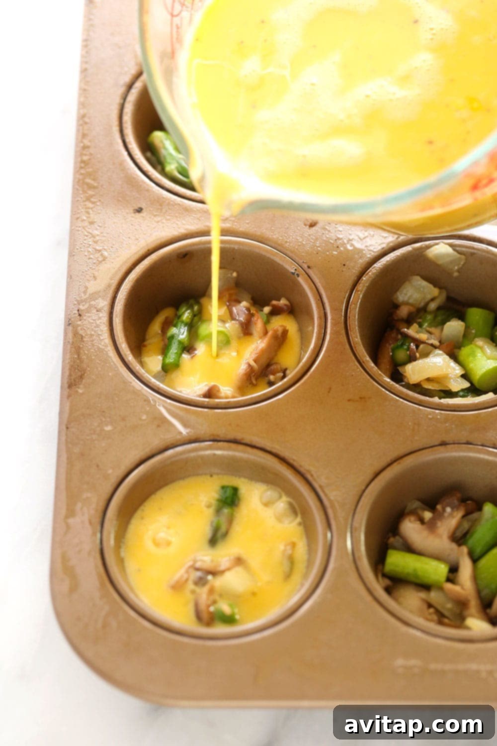 Prepare your healthy asparagus and mushroom egg cups by pouring the mixture into a muffin tin. Hands pouring whisked egg mixture, infused with vegetables, into a muffin tin prepared with non-stick spray.