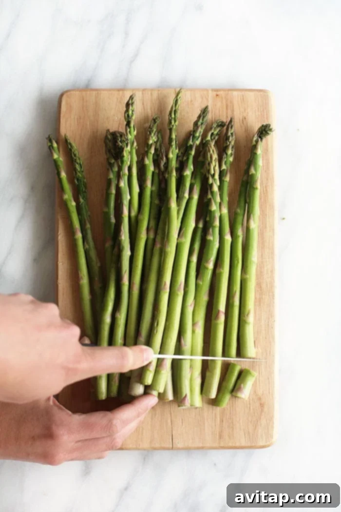 A chef's knife precisely cutting the woody ends off a bundle of fresh green asparagus spears on a wooden cutting board.