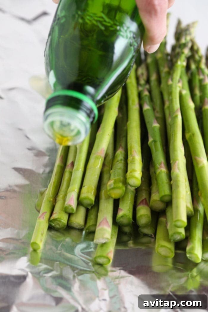 A hand drizzling olive oil generously over a bed of fresh asparagus spears on a large piece of tin foil, ready for seasoning.