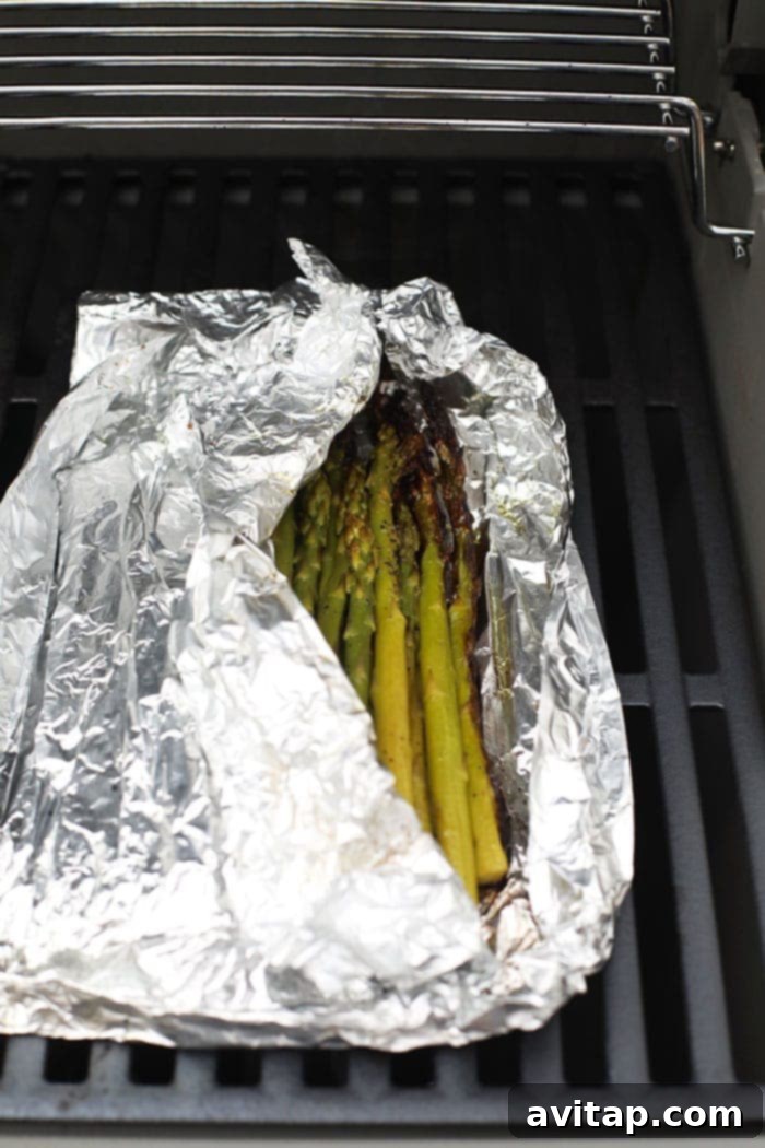A foil pack of seasoned asparagus cooking directly on the hot grates of a barbecue grill, smoke gently rising.