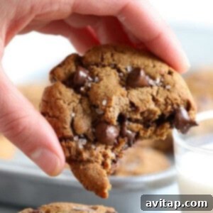 A hand holding a paleo chocolate chip cookie with a bite taken out, next to a glass of milk, emphasizing its deliciousness.