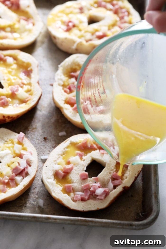 Hands carefully pouring whisked egg mixture into hollowed-out bagels on a baking sheet
