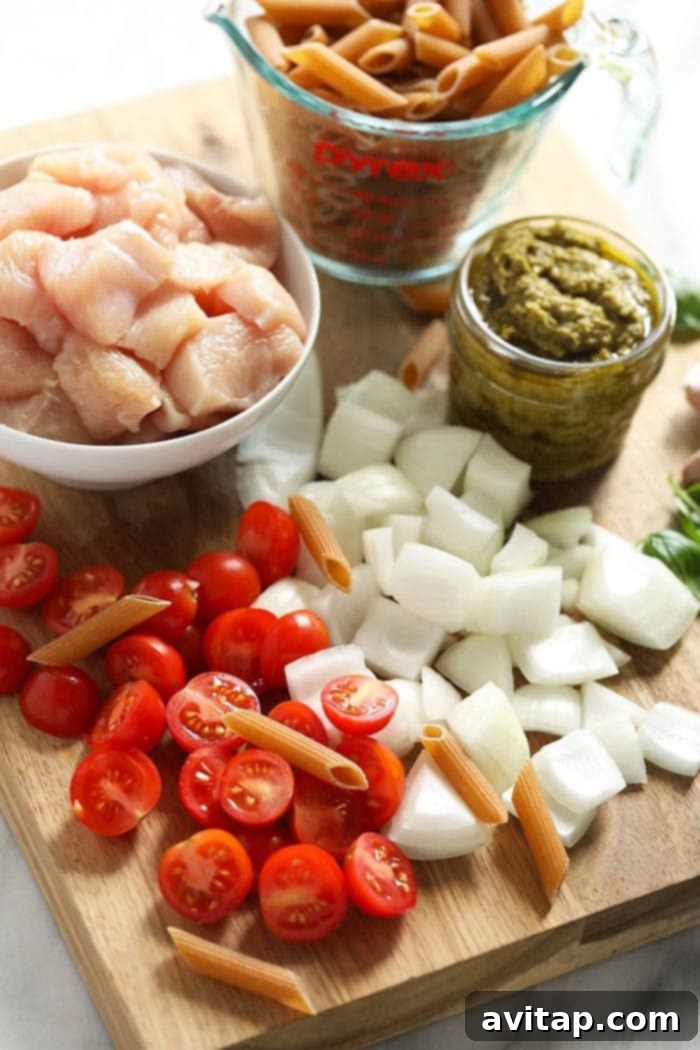 A selection of fresh ingredients laid out, including penne pasta, chicken breast, cherry tomatoes, and basil, ready for making pesto chicken pasta.