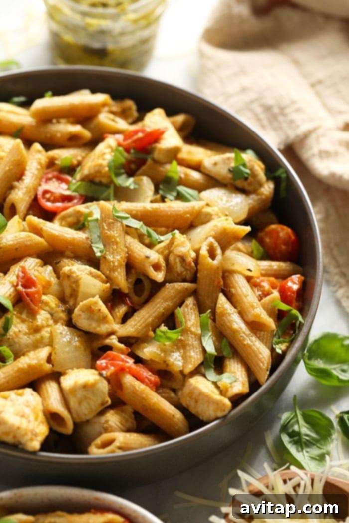 A close-up shot of a bowl of pesto chicken pasta, highlighting the textures of the pasta, chicken, and fresh basil garnish.