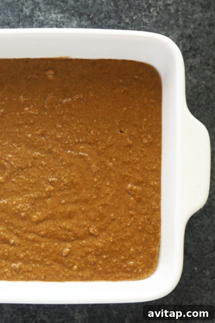 Gingerbread batter evenly spread in a square baking pan, ready to be placed into the oven, showcasing a smooth, rich brown surface.