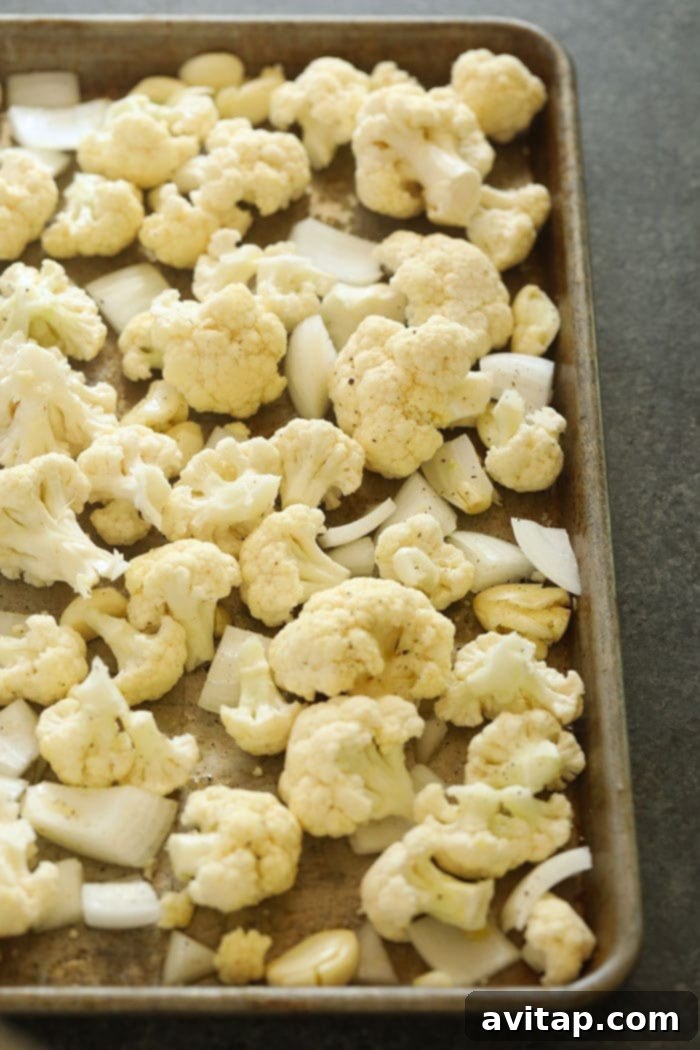 Cauliflower florets, sliced onion, and whole garlic cloves on a baking sheet, prepped for roasting.