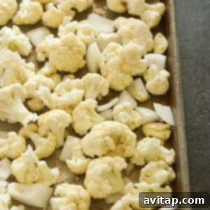 Cauliflower florets, onion, and garlic on a baking sheet, seasoned and ready for roasting.