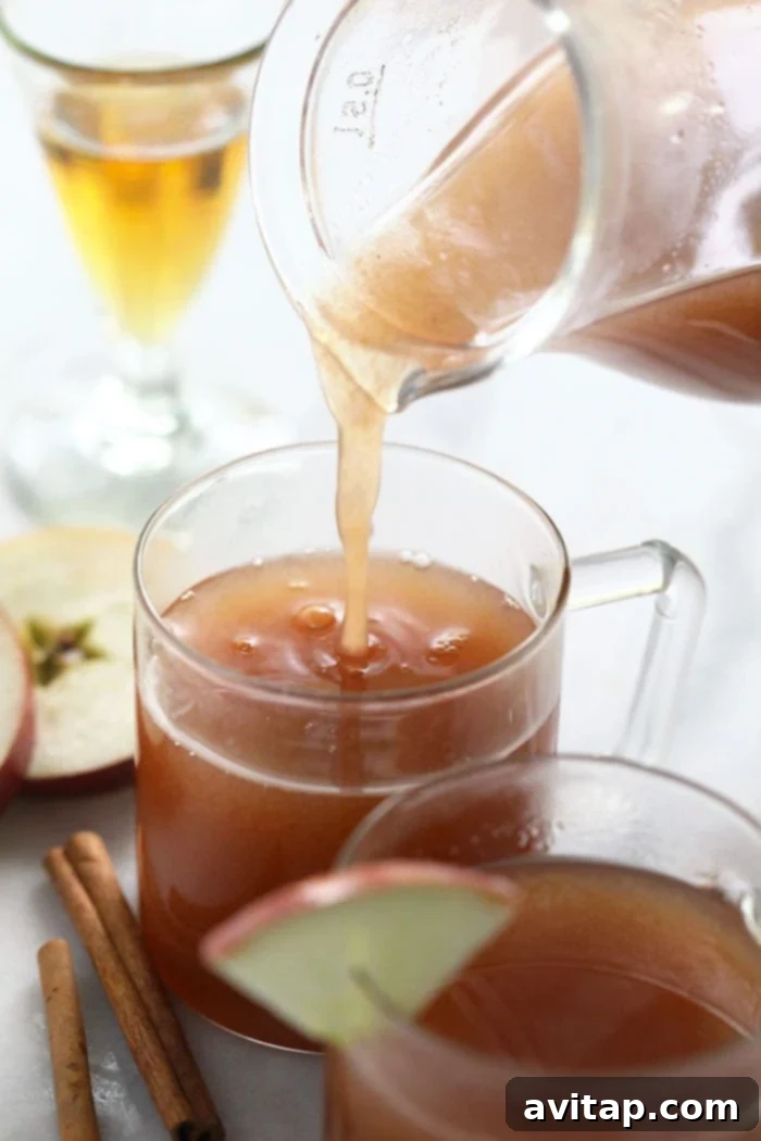 Spiked apple cider being poured into a mug, with fresh apple slices and cinnamon sticks in the background.