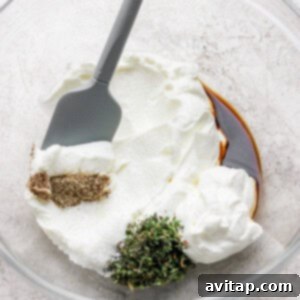 A mixing bowl with creamy yogurt, savory Worcestershire sauce, fresh herbs, and various spices, being stirred with a gray spatula to create the dip's flavorful base.