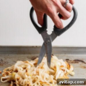 A hand using kitchen scissors to cut cooked onions directly on a baking sheet, preparing them for blending into the creamy dip.