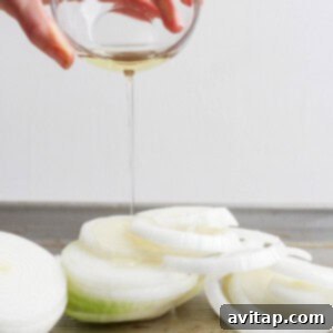A hand pours olive oil from a glass bowl onto sliced onions on a baking sheet, preparing them for roasting.