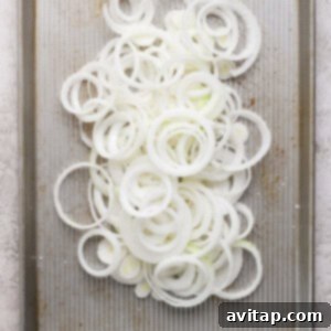 Thinly sliced onion rings spread on a baking sheet with a textured surface, showcasing their even distribution for caramelization.