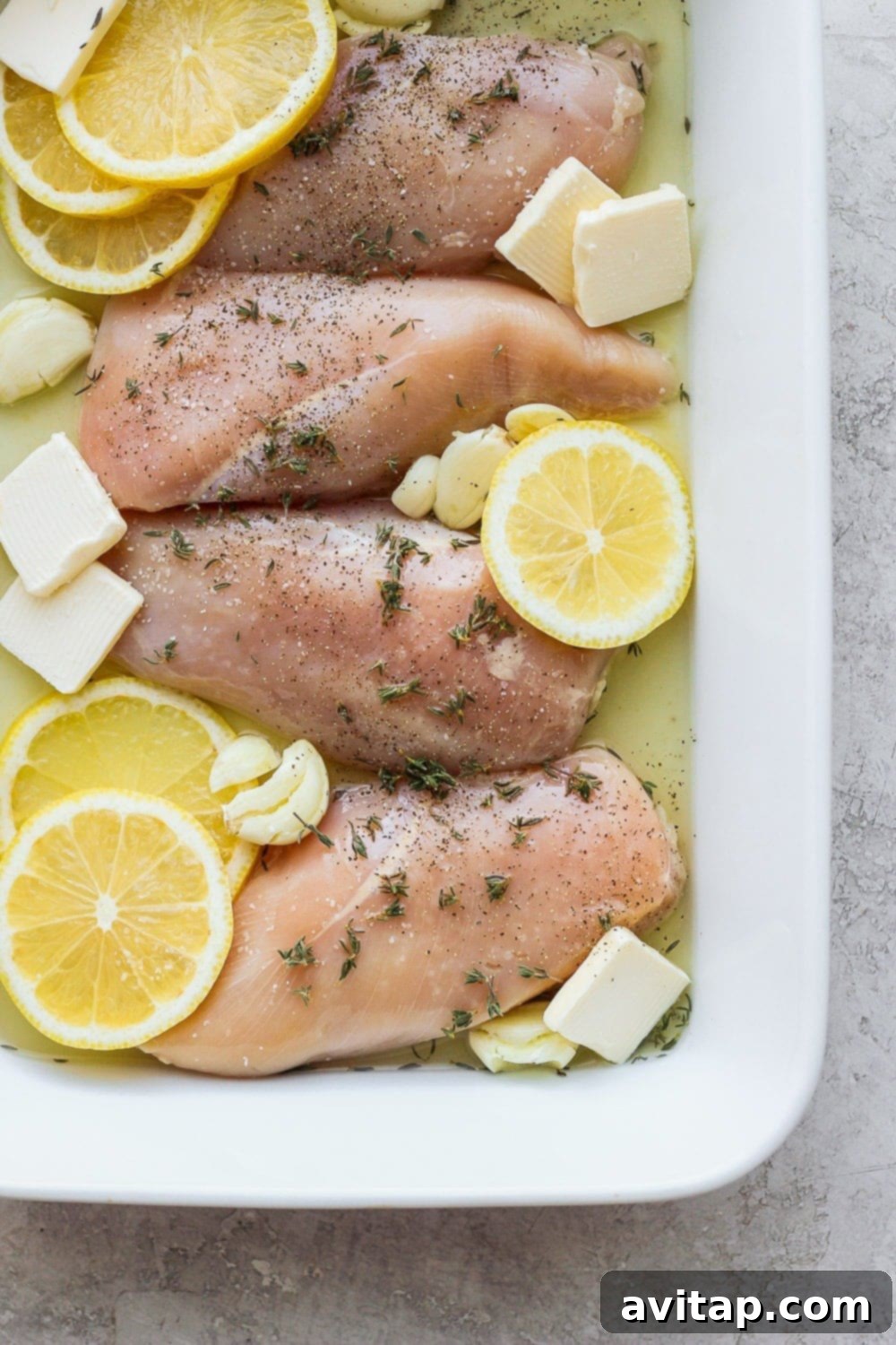Prepare your healthy baked lemon chicken with fresh ingredients for meal prepping throughout the week. Raw chicken breasts nestled with fresh lemon slices, garlic cloves, and pats of butter in a baking dish, pre-marinade.