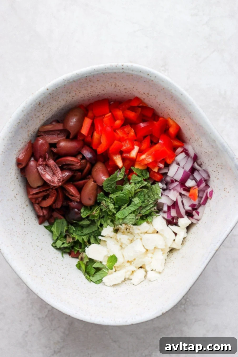 Assortment of fresh Greek salad ingredients, including drained cucumbers and tomatoes, red onion, bell pepper, feta, olives, and mint, in a large bowl, pre-dressing