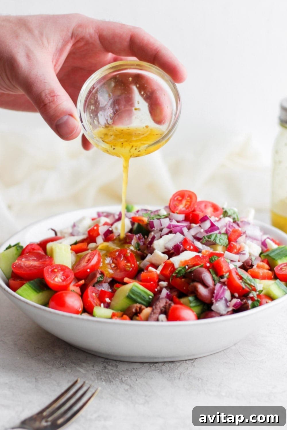 Pouring homemade Greek dressing over a fresh Greek salad in a large, clear bowl, highlighting the colorful ingredients