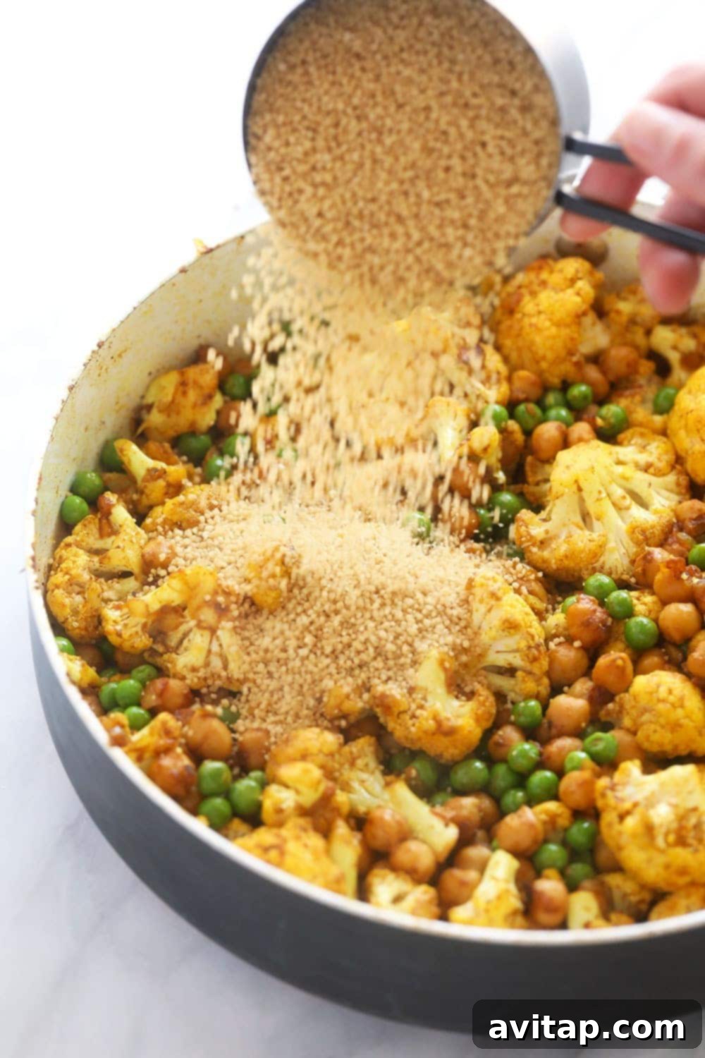 Couscous joining the Moroccan chickpea and vegetable mix in the skillet. Uncooked couscous being poured over the sautéed Moroccan chickpea and vegetable mixture in a skillet.