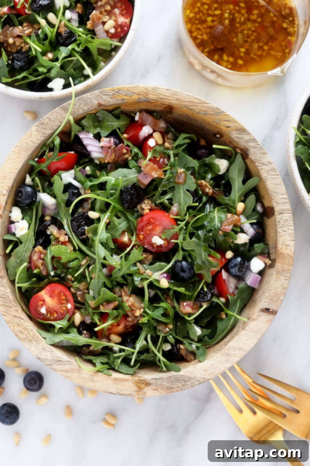 A fresh arugula salad in a mixing bowl, featuring colorful ingredients like blueberries, cherry tomatoes, and pine nuts.