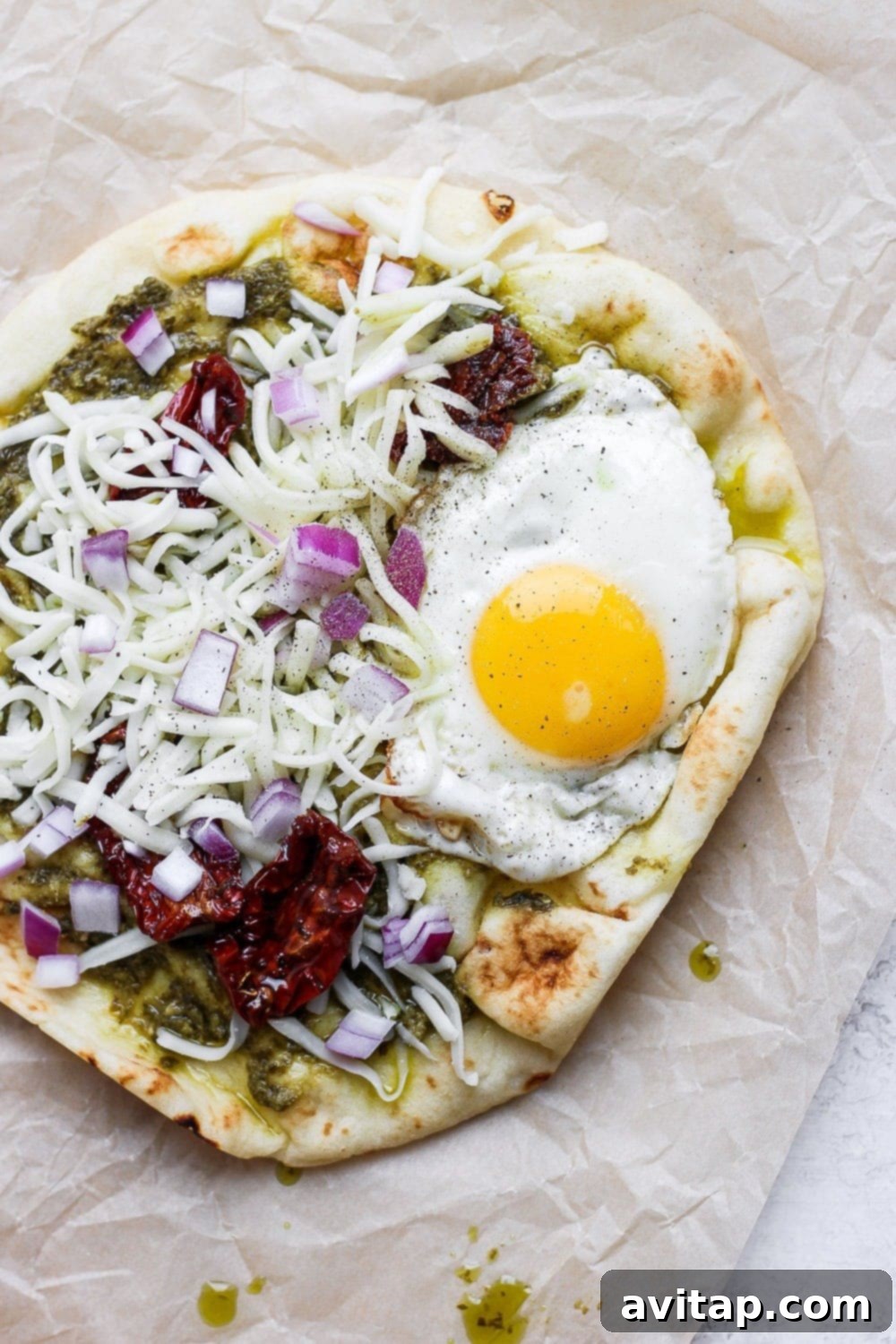 Morning Naan Pizza 5 A fully assembled and baked Naan pizza, adorned with pesto, sun-dried tomatoes, melted cheese, and a perfectly fried egg, ready to be enjoyed.