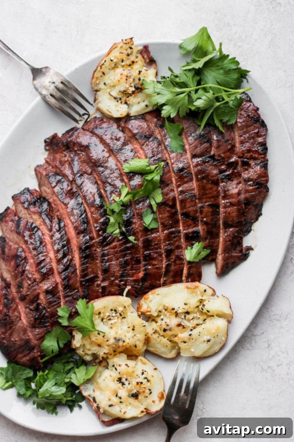 Add this grilled flank steak recipe to the menu this summer! It's tenderized, marinated, and grilled, making this easy grilled flank steak so tender and delicious. Grilled flank steak on a plate with smashed potatoes and fresh parsley, ready to be served.