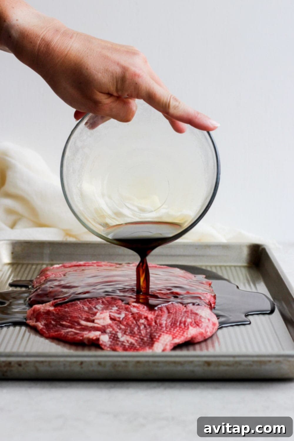 For a more flavorful grilled flank steak, marinate the meat in a delicious soy sauce marinade! You won't regret it! Marinade being poured over a flank steak on a baking sheet, ensuring full coverage and flavor absorption.
