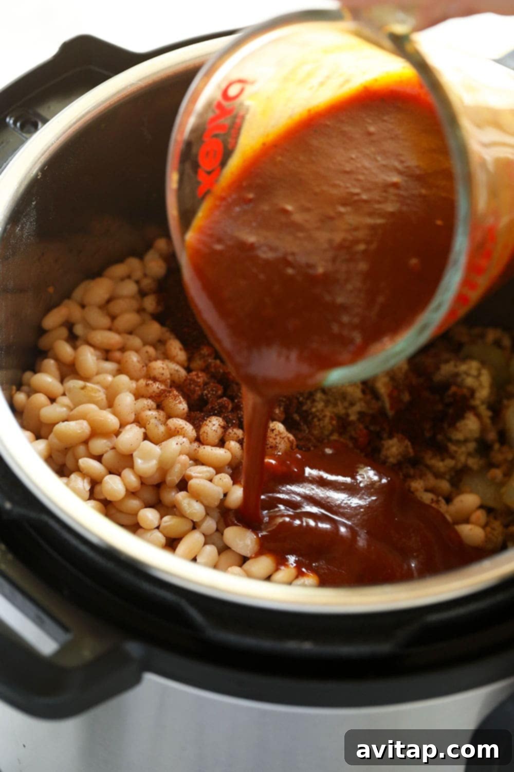 Homemade BBQ sauce being added to Instant Pot baked beans for perfect flavoring. A hand pouring flavorful BBQ sauce into an Instant Pot filled with cooked ingredients.