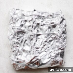 Grilled corn wrapped in aluminum foil on a white surface.