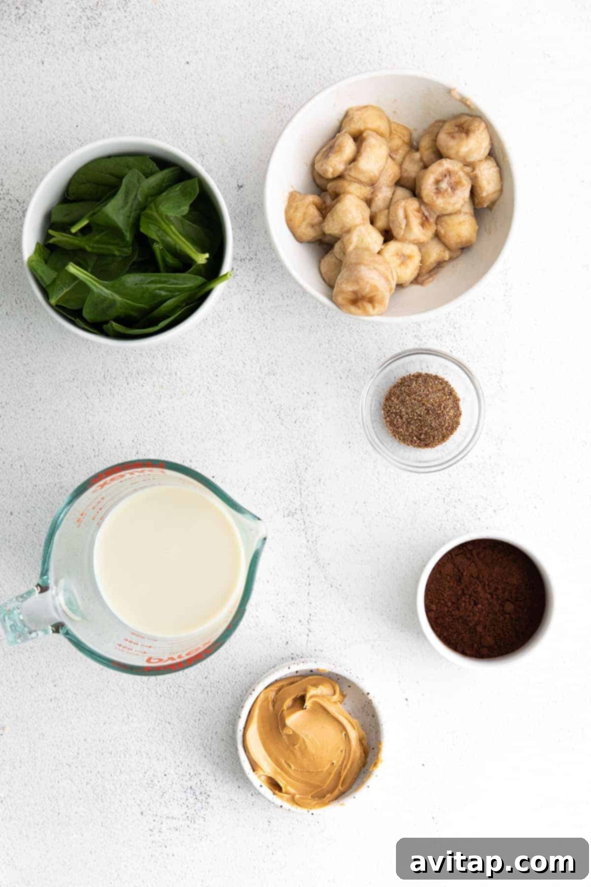 A selection of fresh smoothie ingredients including bananas, spinach, peanut butter, and cocoa powder on a kitchen countertop, ready for blending.