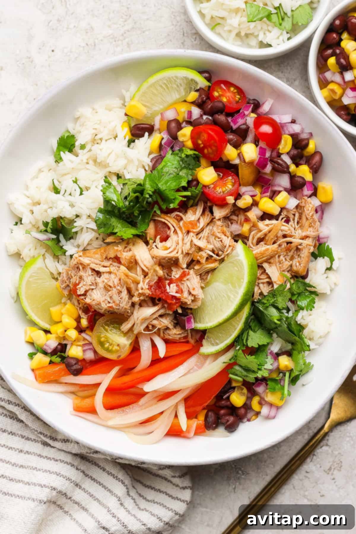 A vibrant bowl of rice topped with perfectly shredded salsa chicken, black beans, corn, diced tomatoes, thinly sliced red onions, a wedge of lime, and fresh cilantro, all beautifully garnished with sliced bell peppers.