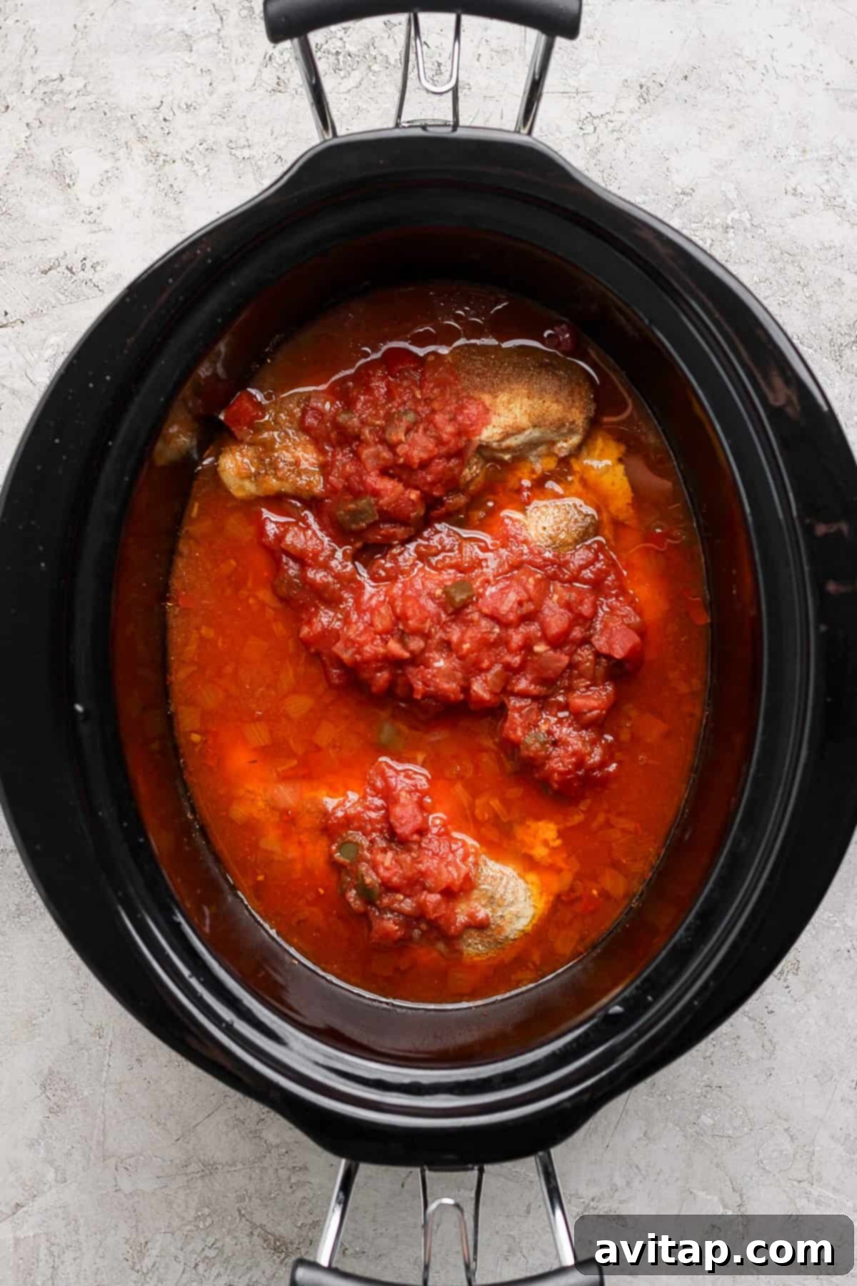 A slow cooker filled with seasoned chicken and rich red tomato salsa, positioned on a bright, minimalist surface.