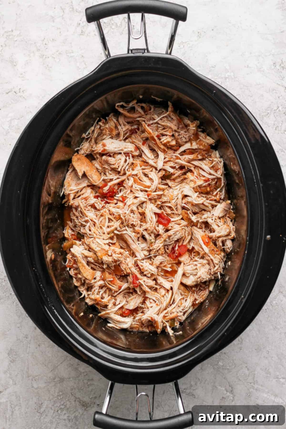 Shredded chicken generously mixed with vibrant red peppers and a rich sauce, waiting in a slow cooker on a modern gray textured surface.