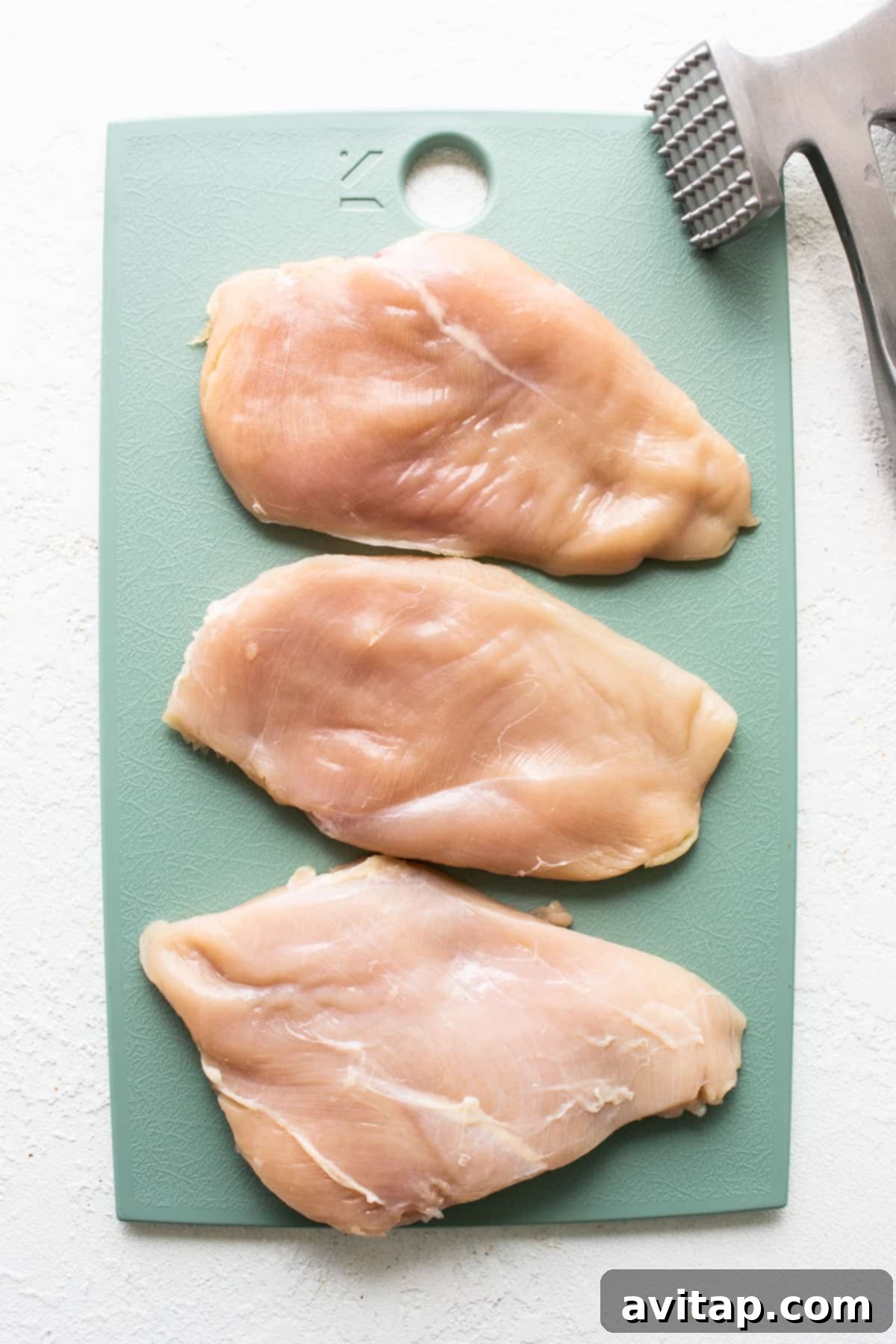 Succulent Grilled Chicken 3 Raw chicken breast being pounded with a meat tenderizer on a wooden cutting board, aiming for an even thickness.