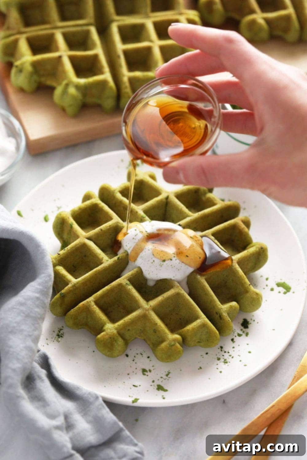 A stack of vibrant green gluten-free matcha waffles with maple syrup being poured over them, topped with fresh berries.