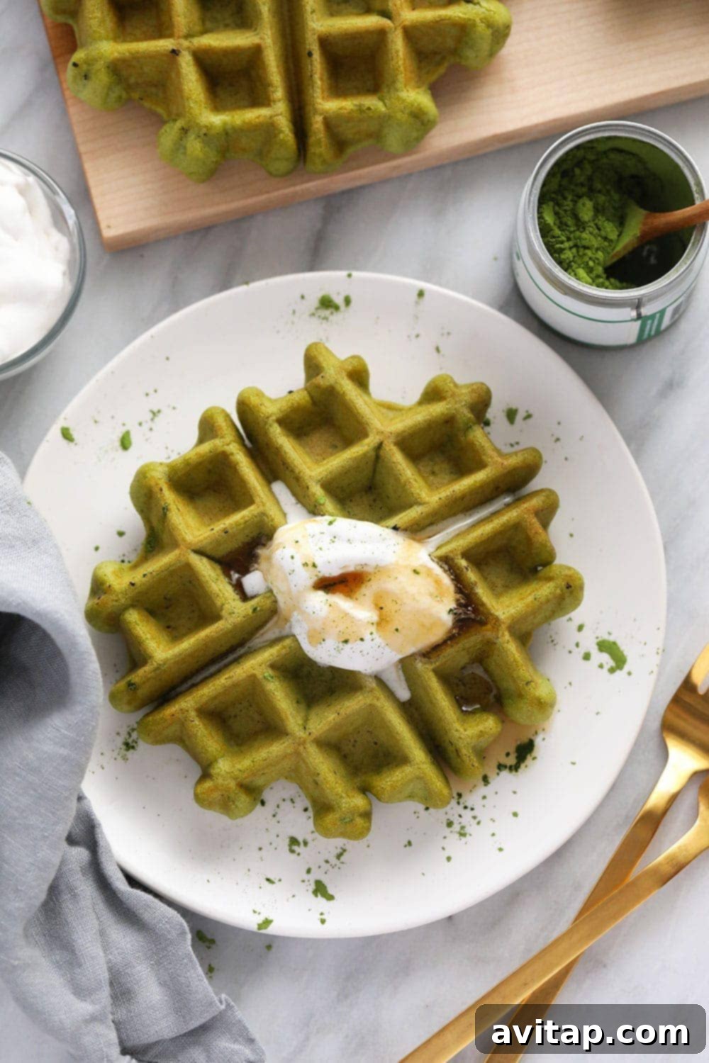 A single green matcha waffle on a white plate, topped with a dollop of coconut whipped cream and a drizzle of maple syrup.