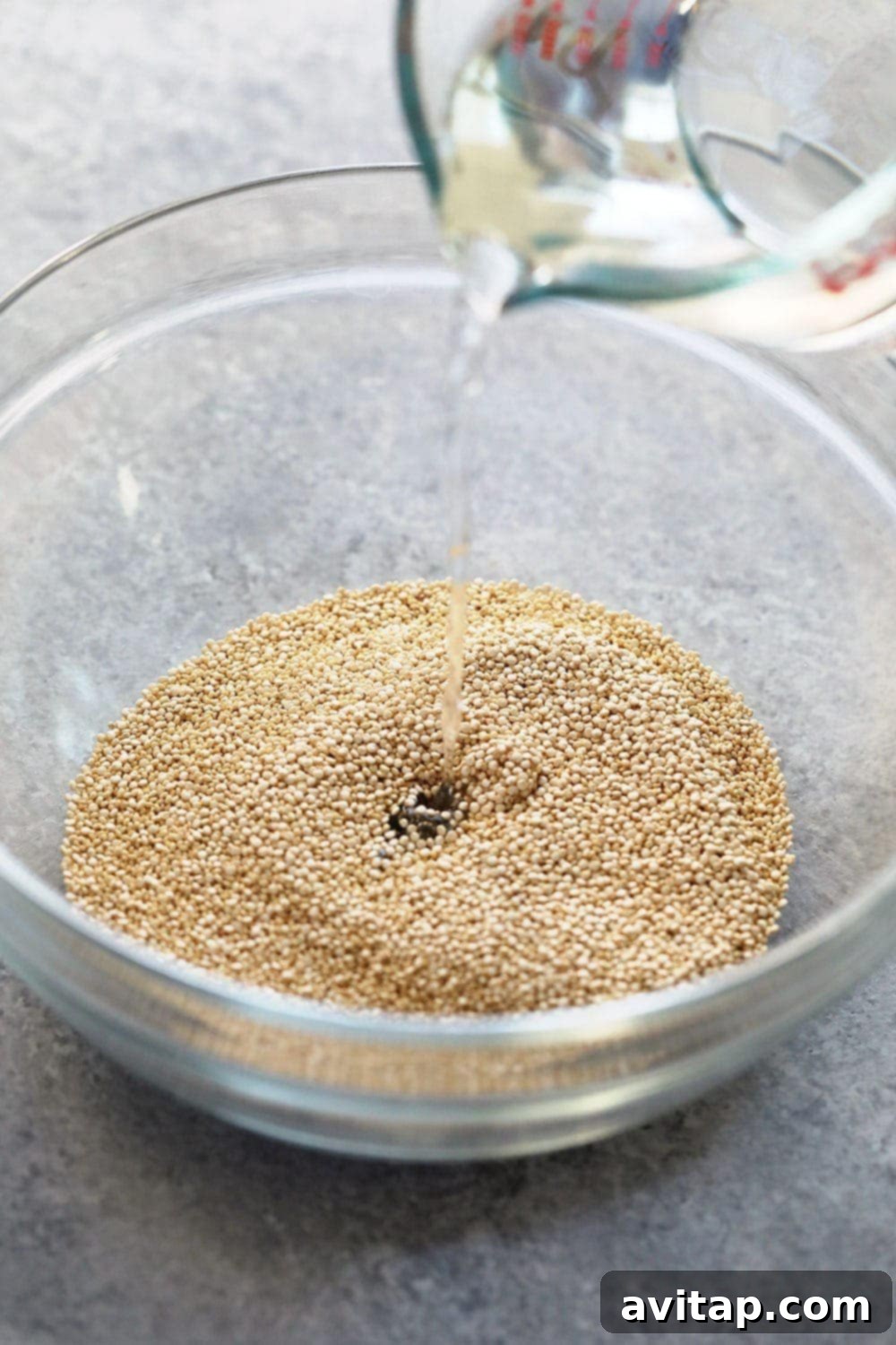 Uncooked white quinoa in a deep, microwave-safe bowl with water being poured over it, illustrating the preparation step.