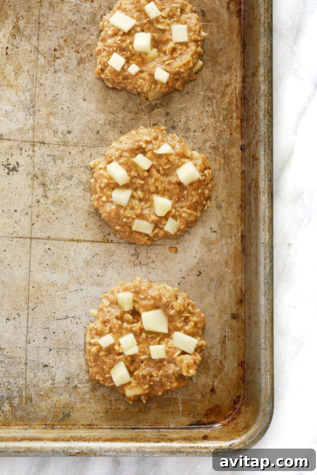 Unbaked apple oatmeal cookies neatly arranged on a baking sheet, topped with fresh apple pieces.