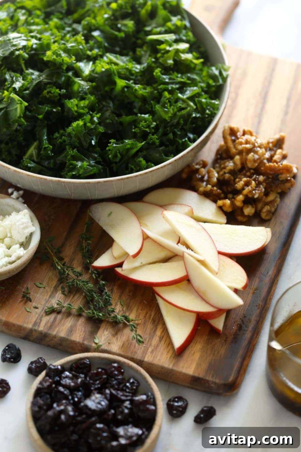 Looking for a last-minute Thanksgiving salad idea? Try our delicious massaged kale, apple, and walnut salad made with goat cheese, dried cherries, and our favorite vinaigrette! A collection of fresh Thanksgiving salad ingredients, including kale, apples, walnuts, and herbs, arranged on a rustic cutting board.