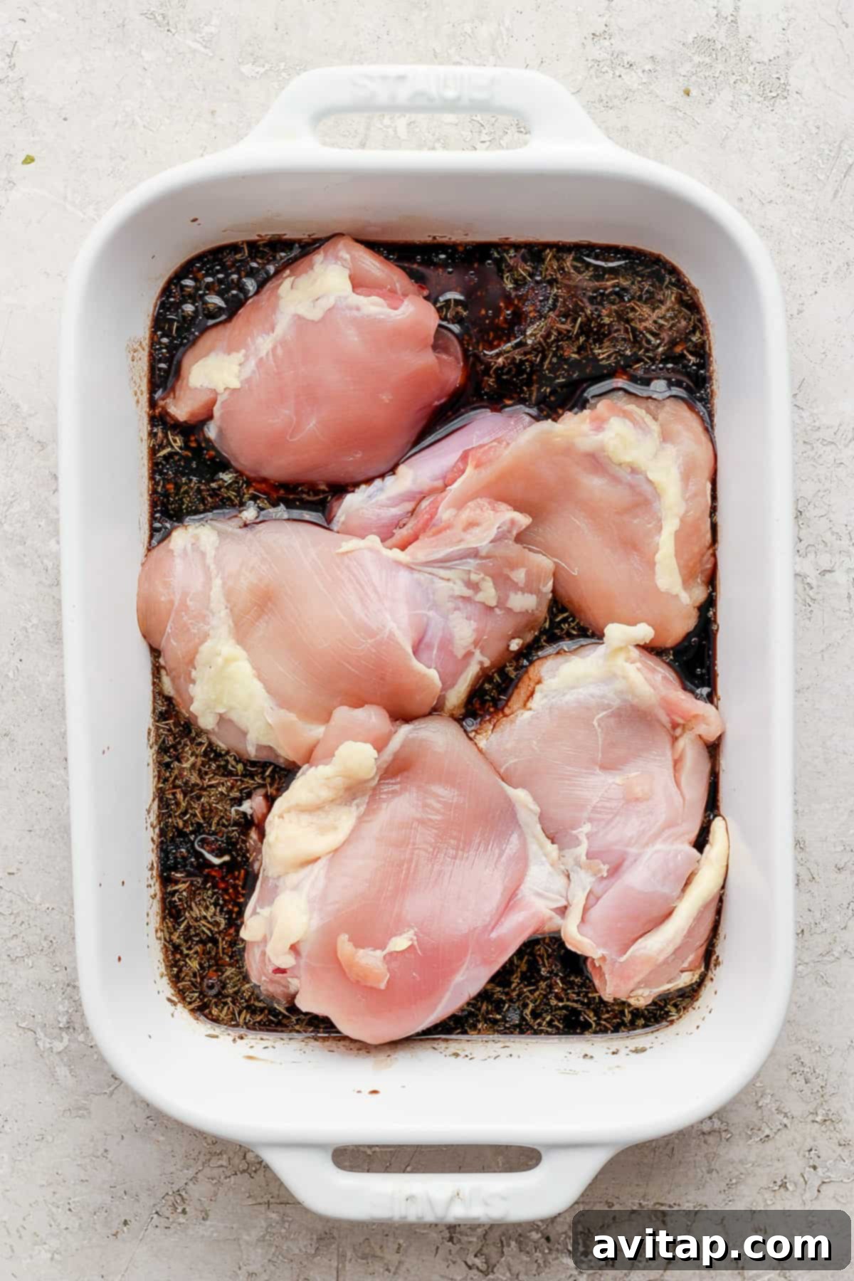 Raw chicken breasts submerged in a dark marinade within a white baking dish.